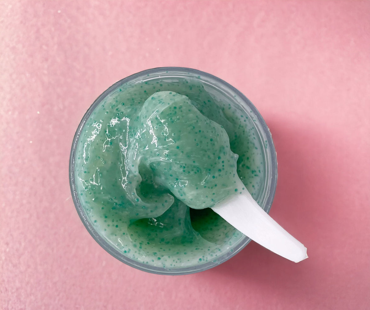 Jar of green jojoba bead face scrub with a spatula on pink