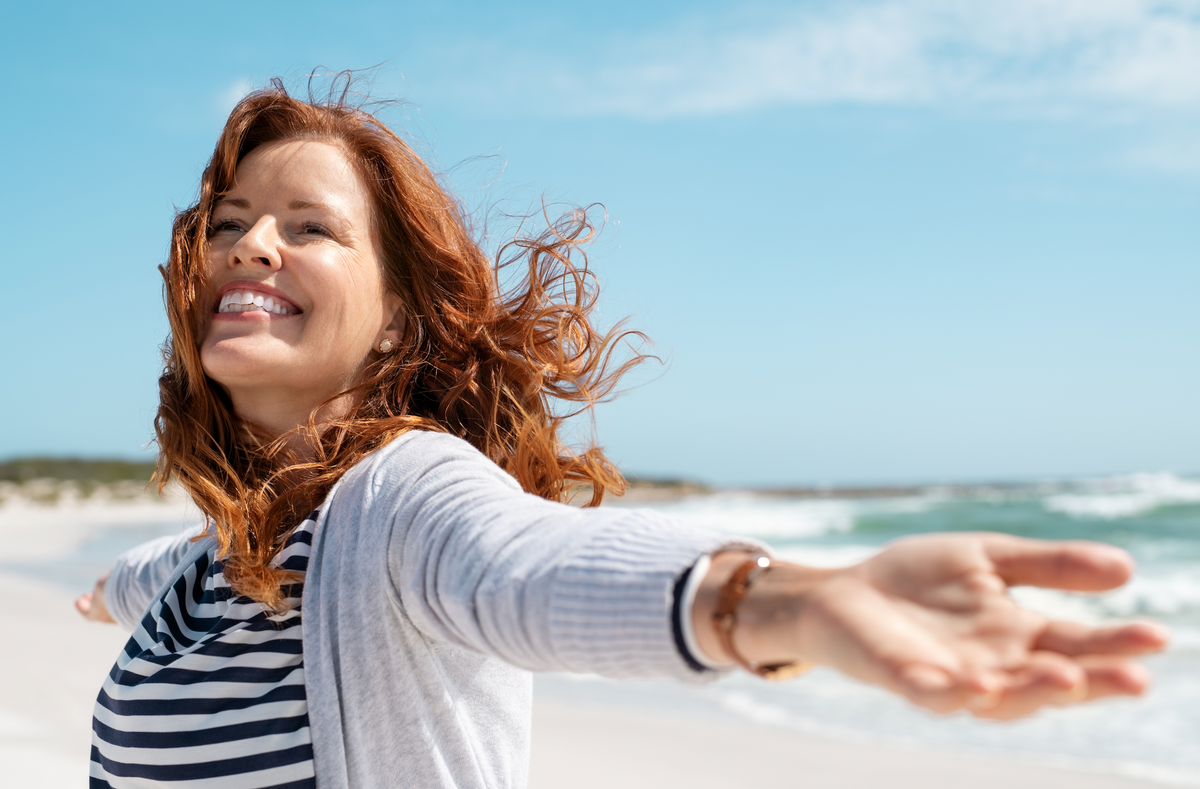 Woman with red hair smiling with arms outstretched on a beach