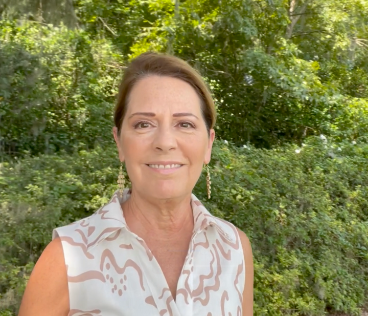 The founder of Magnolia Skin Essentials smiling outdoors in a sleeveless cream blouse with a rose-colored print and gold drop earrings, with green garden foliage behind her